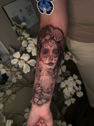 Intricate black and grey tattoo of a sugar skull woman with butterflies and roses on forearm.