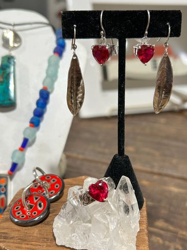 Handcrafted jewelry with red heart stones and feather designs displayed on crystal and velvet stands.