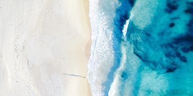 Aerial view of waves meeting a sandy beach with a lone person casting a long shadow.