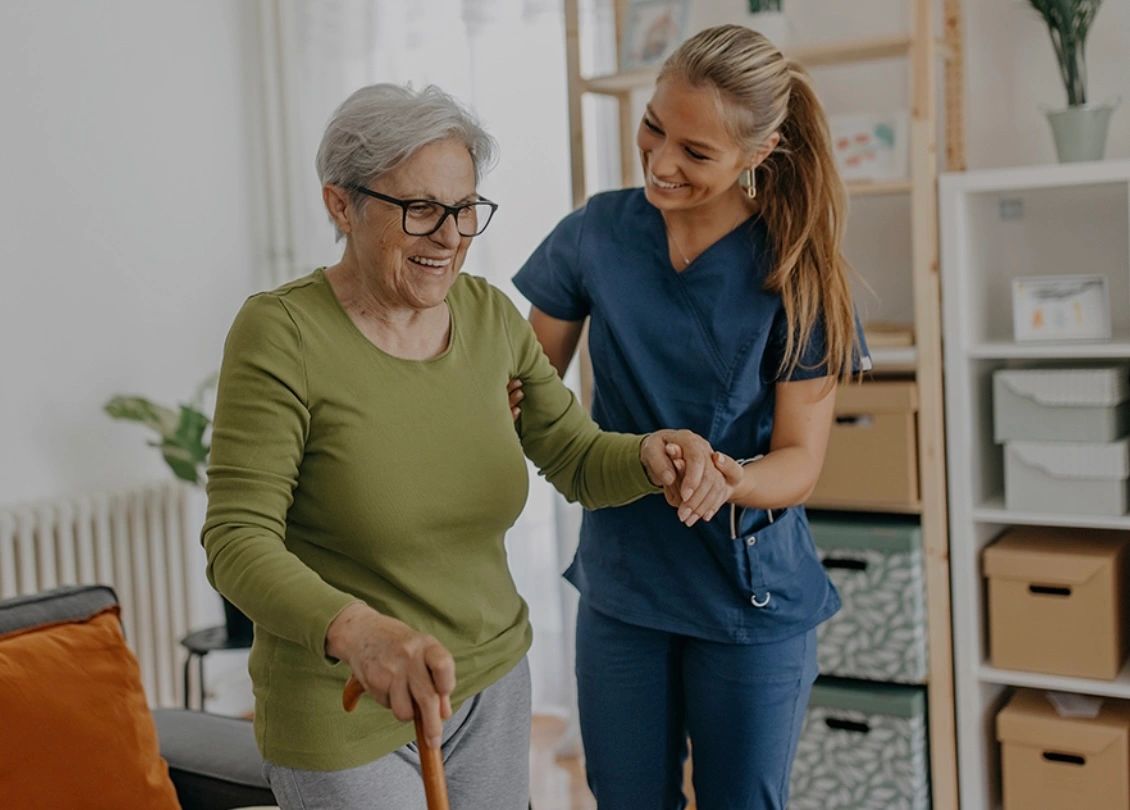 A nurse assists an elderly woman with walking, both smiling warmly.