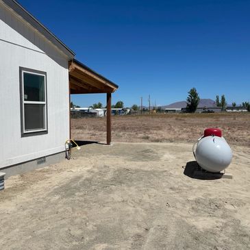 A white house with a propane tank on a dry plot of land under a clear blue sky.