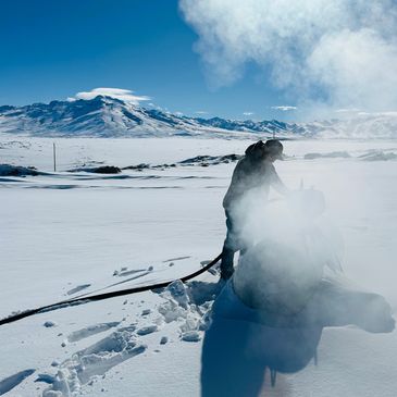 A person working with equipment in a snowy, mountainous landscape with steam rising.