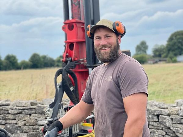 Smiling man in work gear stands outdoors near machinery and stone wall.