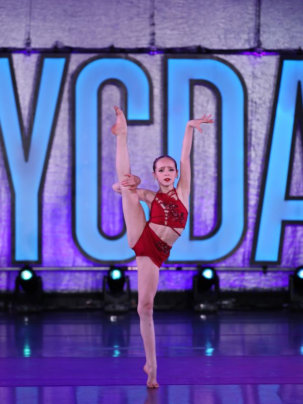 Young dancer performing a high leg hold on stage in a red costume.