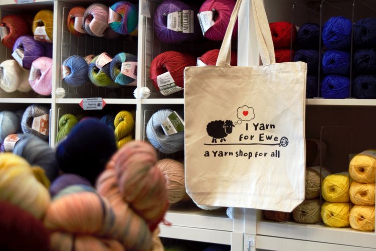 A tote bag featuring the I Yarn For Ewe logo in front of a wall of yarn.