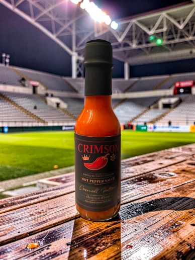 A bottle of Crimson hot pepper sauce on a wooden table in a stadium.
