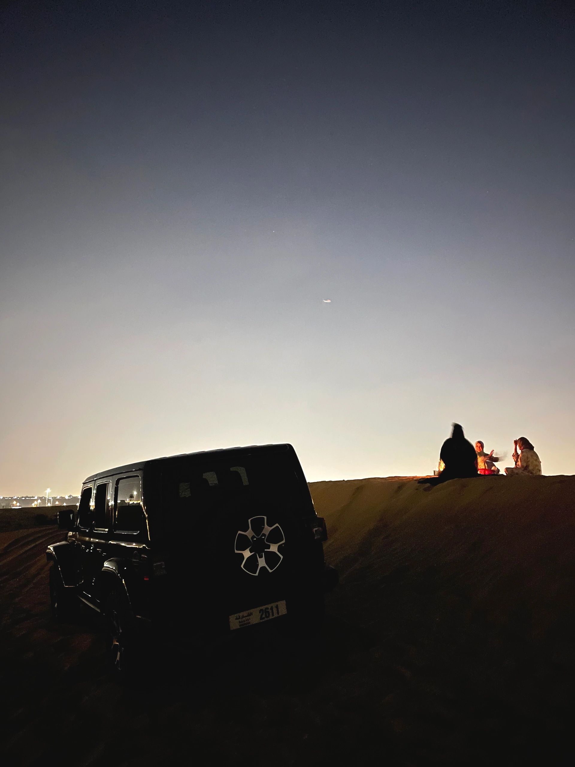 Nighttime desert scene with a parked jeep and three people sitting on a dune.