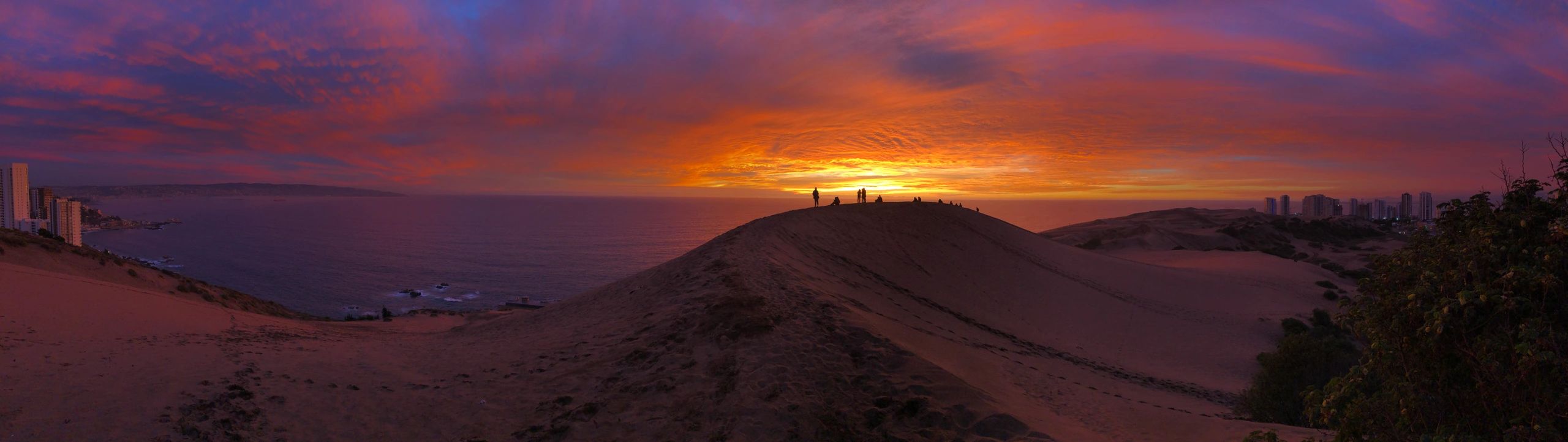 Sunset over sand dunes with silhouetted people and coastal cityscape.