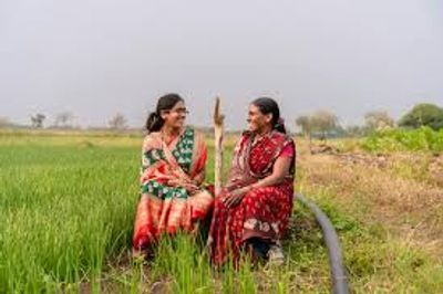 Two women in colorful sarees sitting and chatting in a green field.