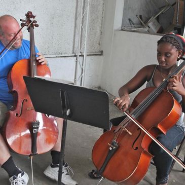 Two people playing cellos together, one smiling and the other focused.