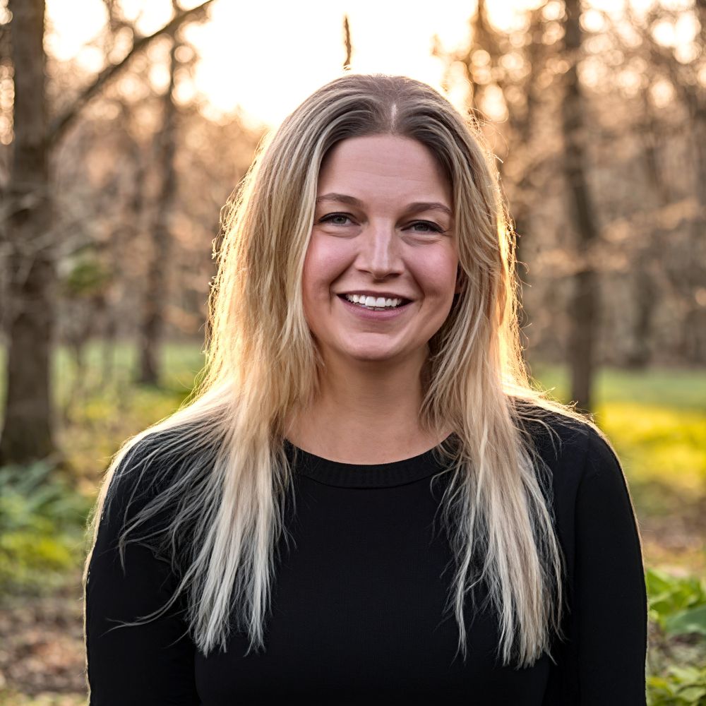Smiling woman with long blonde hair in a black top outdoors during golden hour.