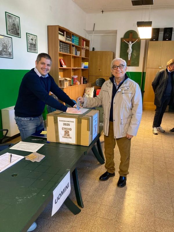Two men smiling near a voting box labeled 2025 elections.