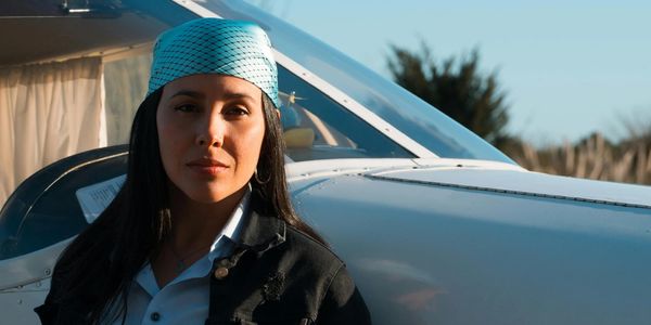 A woman with a blue headscarf stands next to a small airplane under a clear sky.