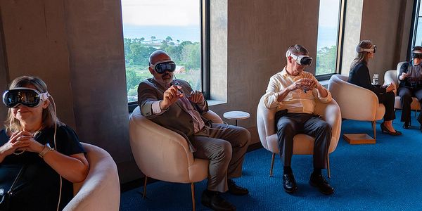 Group of people sitting in chairs wearing virtual reality headsets inside a room with large windows.