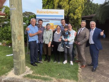 Group of people smiling in front of a Raine & Horne real estate sign.