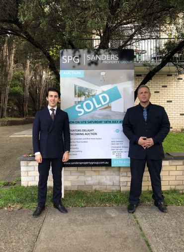 Two men in suits standing beside a sold real estate auction sign.