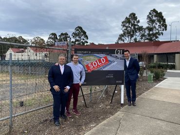 Three men stand next to a sold Century 21 real estate sign in front of a fenced lot.