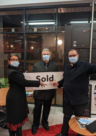 Three masked individuals holding a 'Sold' sign indoors.