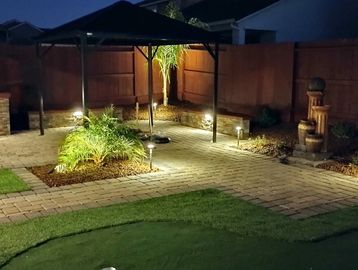 Cozy backyard patio illuminated by soft garden lights and a shaded umbrella at night.