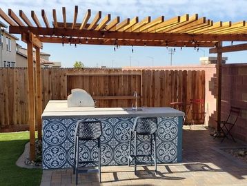 Outdoor kitchen with tiled counter and wooden pergola in backyard.