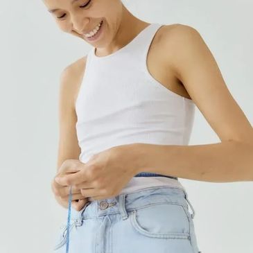 Smiling woman measuring waist with tape measure, wearing white tank top and light jeans.