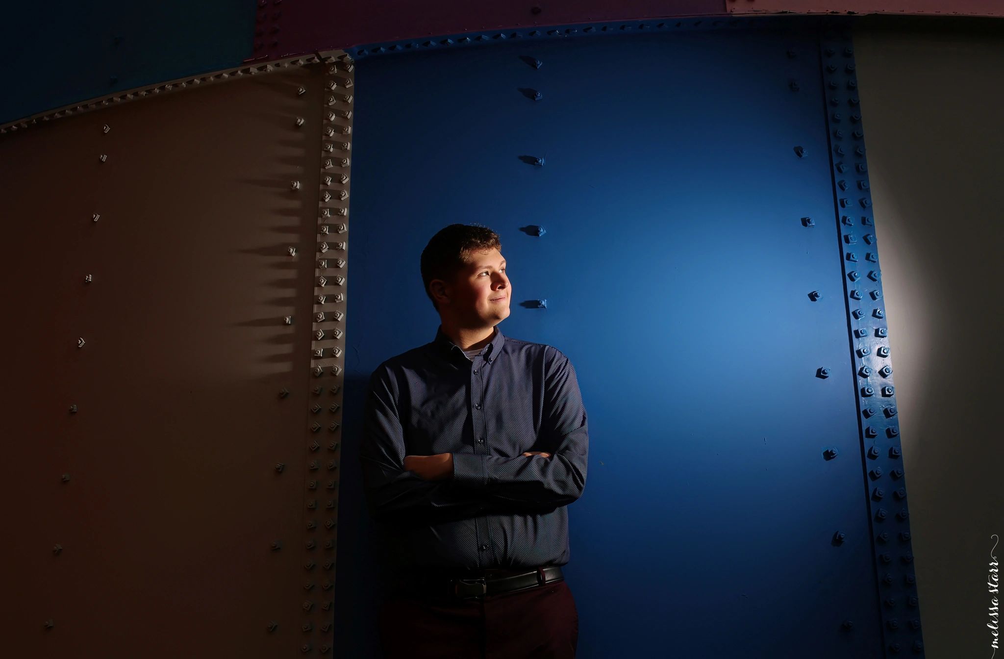 Young man with folded arms gazing to the side against a blue and brown riveted metal wall.