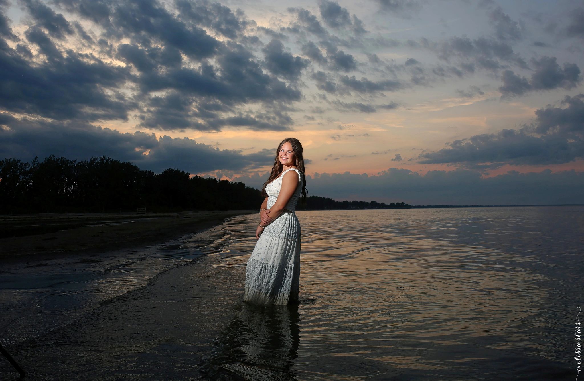 Young woman in a white dress stands in shallow water at sunset.