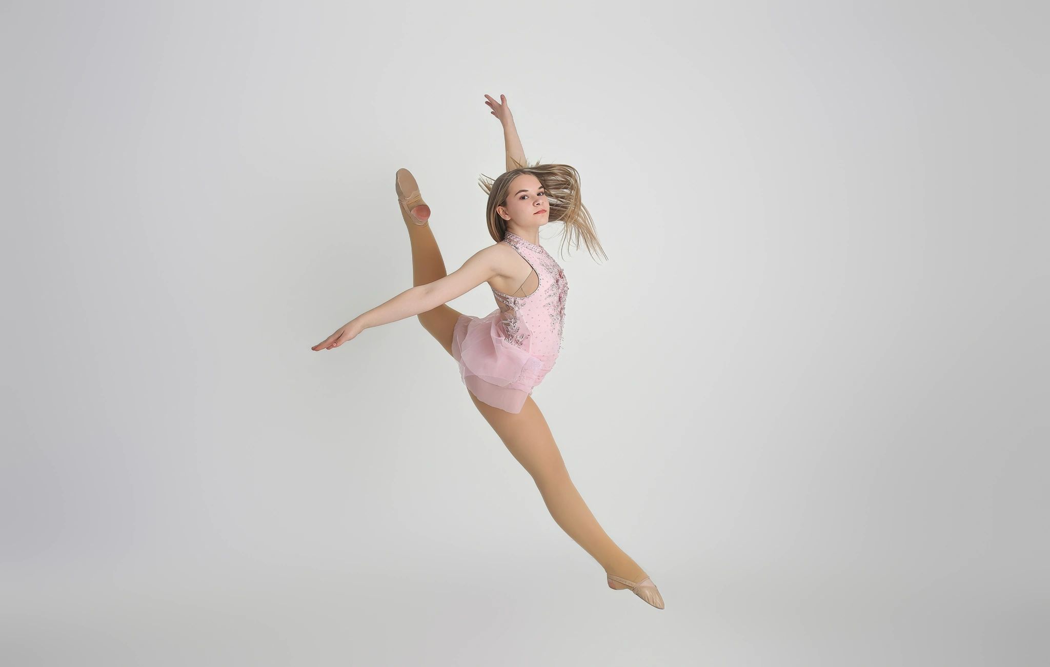 Young ballerina leaps gracefully in a pink costume against a plain background.