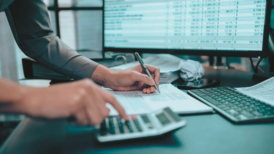 Person calculating finances with calculator and pen near a computer.