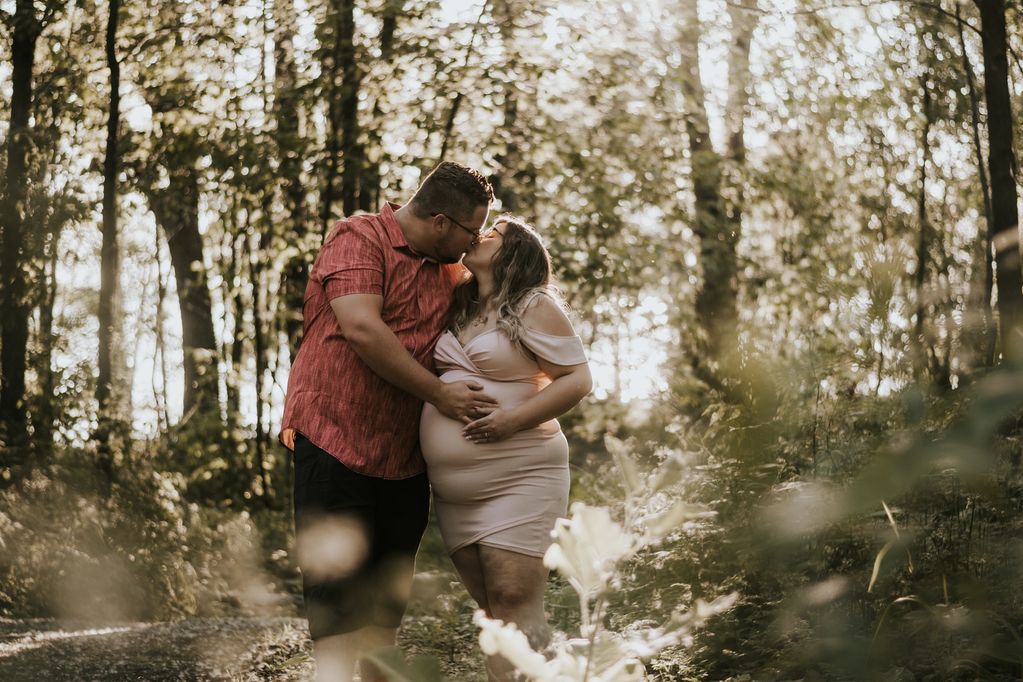 Un jeune couple enceinte, mains sur le ventre, s'embrasse dans la forêt.