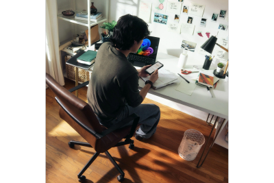 Person working at a desk with a laptop and phone in a sunlit room.