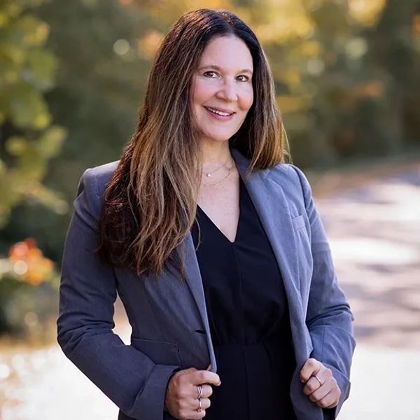 A confident woman in a gray blazer smiles outdoors with a blurred natural background.