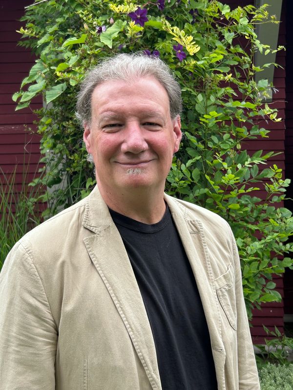 Smiling man in beige blazer standing outdoors with green plants.