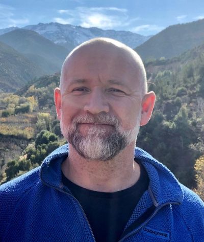 Smiling man in blue jacket with mountainous landscape behind.