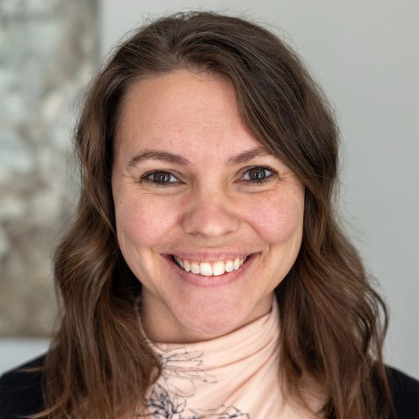A headshot of a woman smiling with brown eyes, long brown hair and a light pink turtleneck shirt