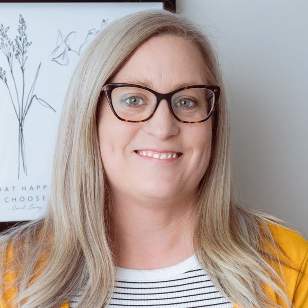 A headshot of a woman smiling with glasses, long blond hair and a striped shirt and a yellow shall