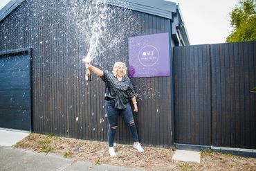 Woman joyfully popping champagne outside a modern building with black fence.
