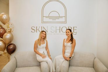 Two women in white outfits sitting on a beige couch at Skin Shore clinic.