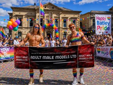 Two muscular men holding a banner at Batley Pride parade with colorful decorations and crowd.
