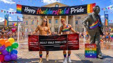 Two men holding a banner at Huddersfield Pride celebration in colorful attire.