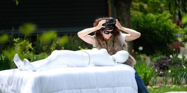A woman adjusts a headpiece while leaning over a mannequin on a white-covered table outdoors.