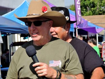 Two men wearing hats, one holding a microphone at an outdoor event.
