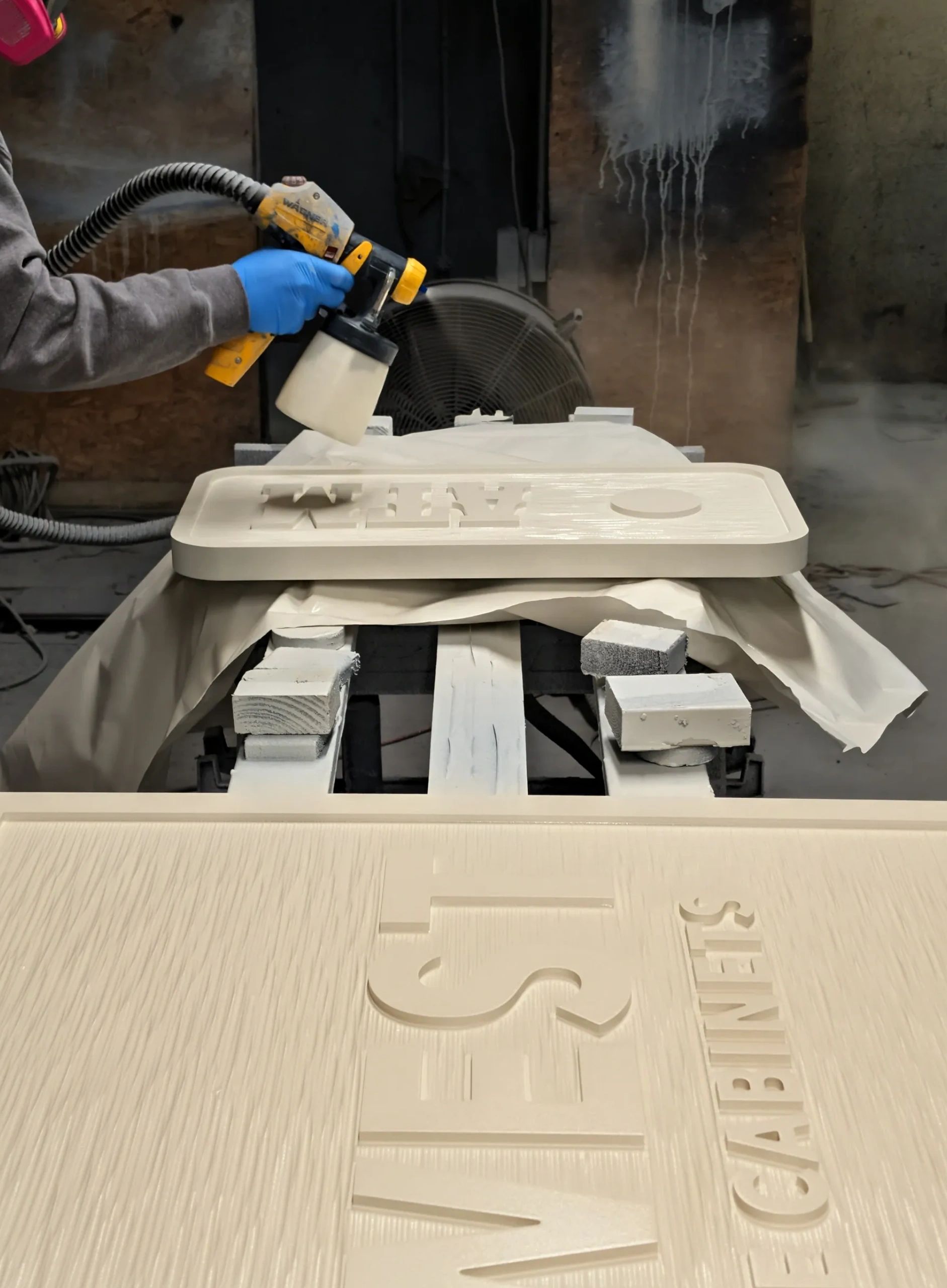 Person spray painting a wooden sign with raised letters in a workshop.