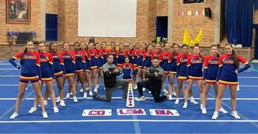 Cheerleading team posing in uniform on a gym mat with coaches kneeling in front.