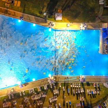 Aerial view of a crowded illuminated swimming pool at night with many people and sun loungers.