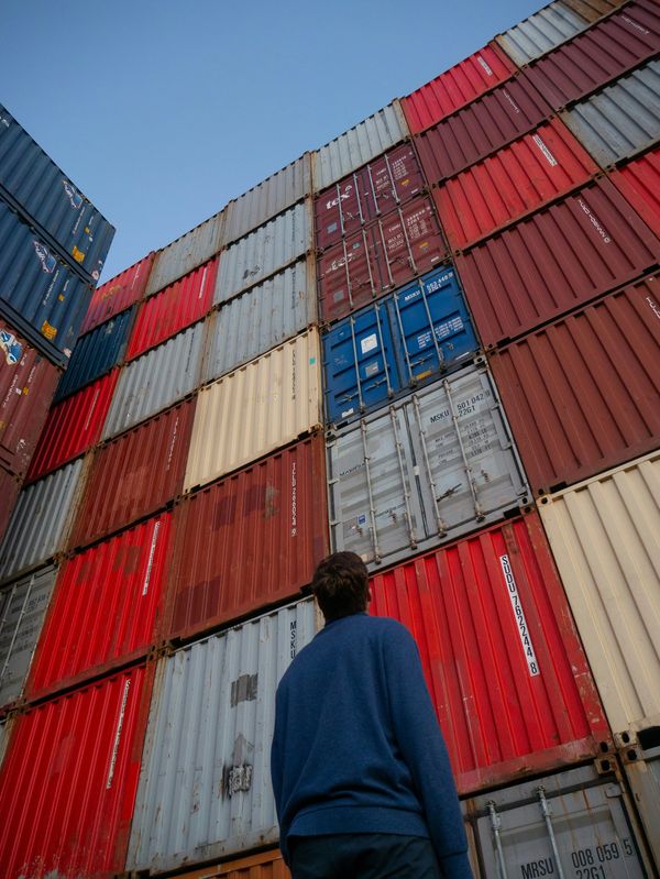 A man in a blue sweater looks up at stacked colorful shipping containers.