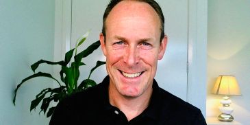 Smiling man with a plant and lamp in the background.