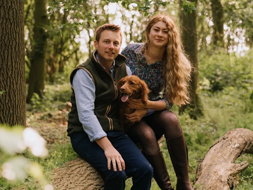 Farmer couple sitting on a log with cocker spaniel in woods.