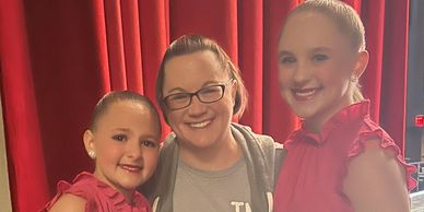 Two young dancers in red dresses with a smiling woman between them in front of red curtains.