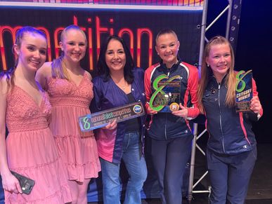 Group of smiling dancers and a woman holding dance competition awards and plaques.